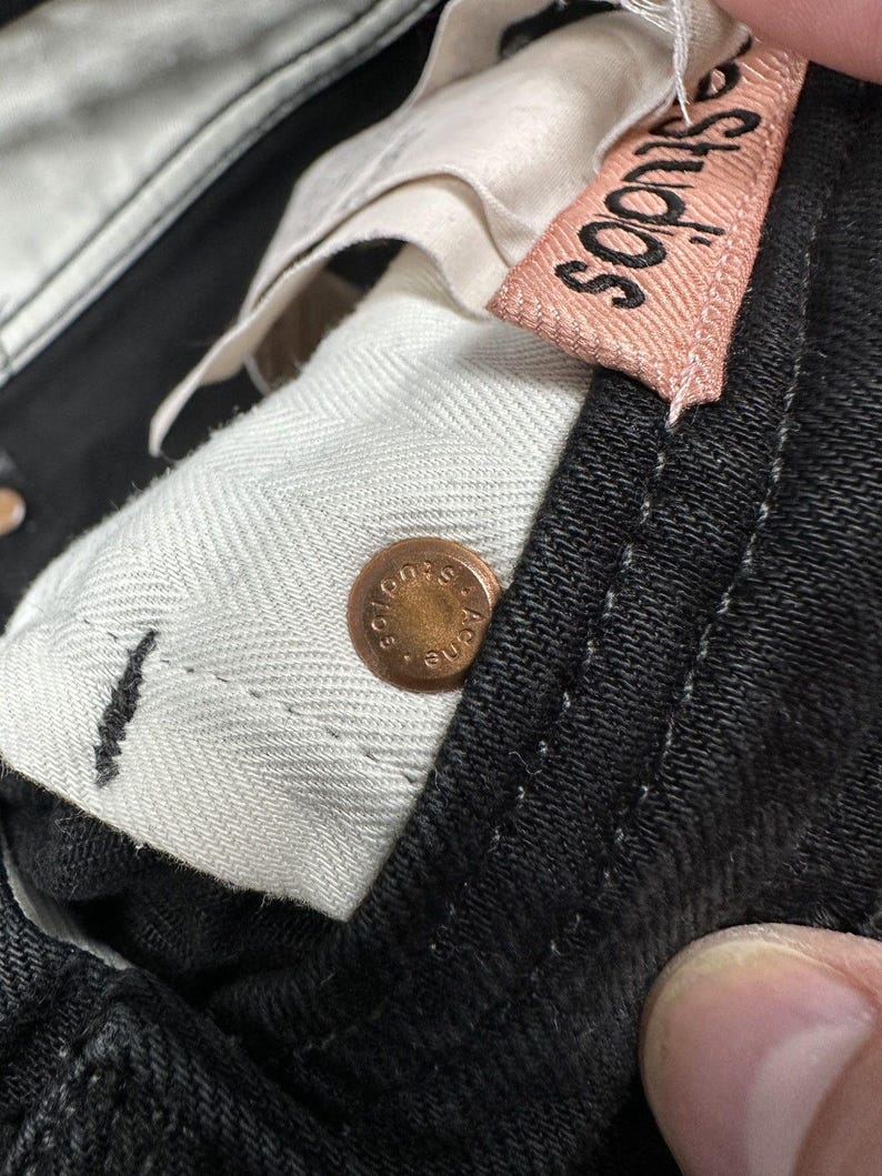 Pode incluir: Cal&ccedil;as de ganga pretas com um rebite de metal cor de cobre gravado com o texto Acne Studios. Uma etiqueta de tecido rosa claro com o texto Acne Studios est&aacute; cosida no c&oacute;s interior, forrado com tecido branco com padr&atilde;o espinha.