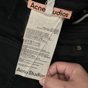 Pode incluir: Cal&ccedil;as de ganga pretas com uma etiqueta de tecido rosa com o texto Acne Studios em preto. Uma etiqueta de cuidados branca est&aacute; anexada, detalhando uma composi&ccedil;&atilde;o de 94% algod&atilde;o, 5% poli&eacute;ster e 1% elastano, com informa&ccedil;&otilde;es da marca.