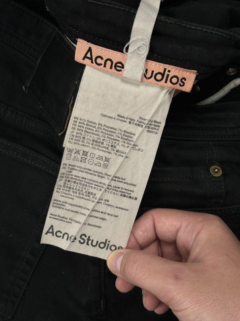 Pode incluir: Cal&ccedil;as de ganga pretas com uma etiqueta de tecido rosa com o texto Acne Studios em preto. Uma etiqueta de cuidados branca est&aacute; anexada, detalhando uma composi&ccedil;&atilde;o de 94% algod&atilde;o, 5% poli&eacute;ster e 1% elastano, com informa&ccedil;&otilde;es da marca.