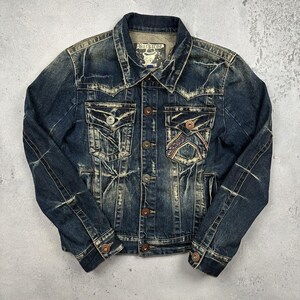May include: A dark blue denim jacket with a distressed, faded wash, featuring a pointed collar, button-front closure, and two chest pockets. One pocket displays decorative stitching. The interior label reads Buckaroo with a hat graphic.