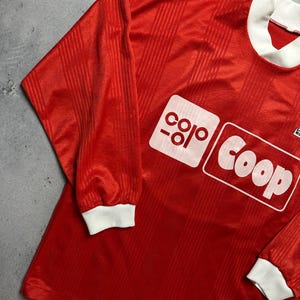 Puede incluir: Camiseta de f&uacute;tbol roja de manga larga con cuello y pu&ntilde;os blancos. La camiseta presenta un logotipo rectangular blanco con la palabra "Coop". La tela tiene un sutil patr&oacute;n de rayas verticales.