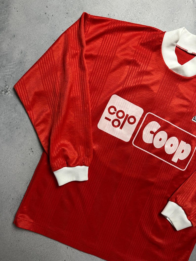 Puede incluir: Camiseta de f&uacute;tbol roja de manga larga con cuello y pu&ntilde;os blancos. La camiseta presenta un logotipo rectangular blanco con la palabra "Coop". La tela tiene un sutil patr&oacute;n de rayas verticales.