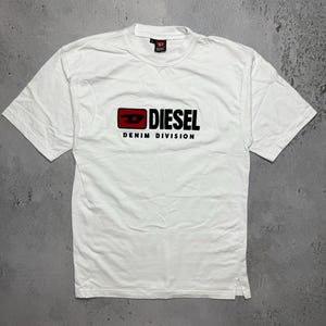 Könnte beinhalten: Weißes T-Shirt mit dem Wort "DIESEL" in schwarzen Buchstaben und einem roten Rechteck-Logo. Darunter steht "DENIM DIVISION" in Schwarz. Das T-Shirt hat kurze Ärmel und einen Rundhalsausschnitt.