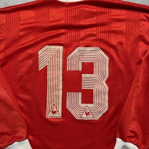 Puede incluir: Camiseta roja de manga larga con el n&uacute;mero 13 en blanco en la espalda. La camiseta tiene cuello y pu&ntilde;os blancos. Un peque&ntilde;o logotipo es visible debajo del n&uacute;mero. La tela tiene un sutil patr&oacute;n de rayas verticales.