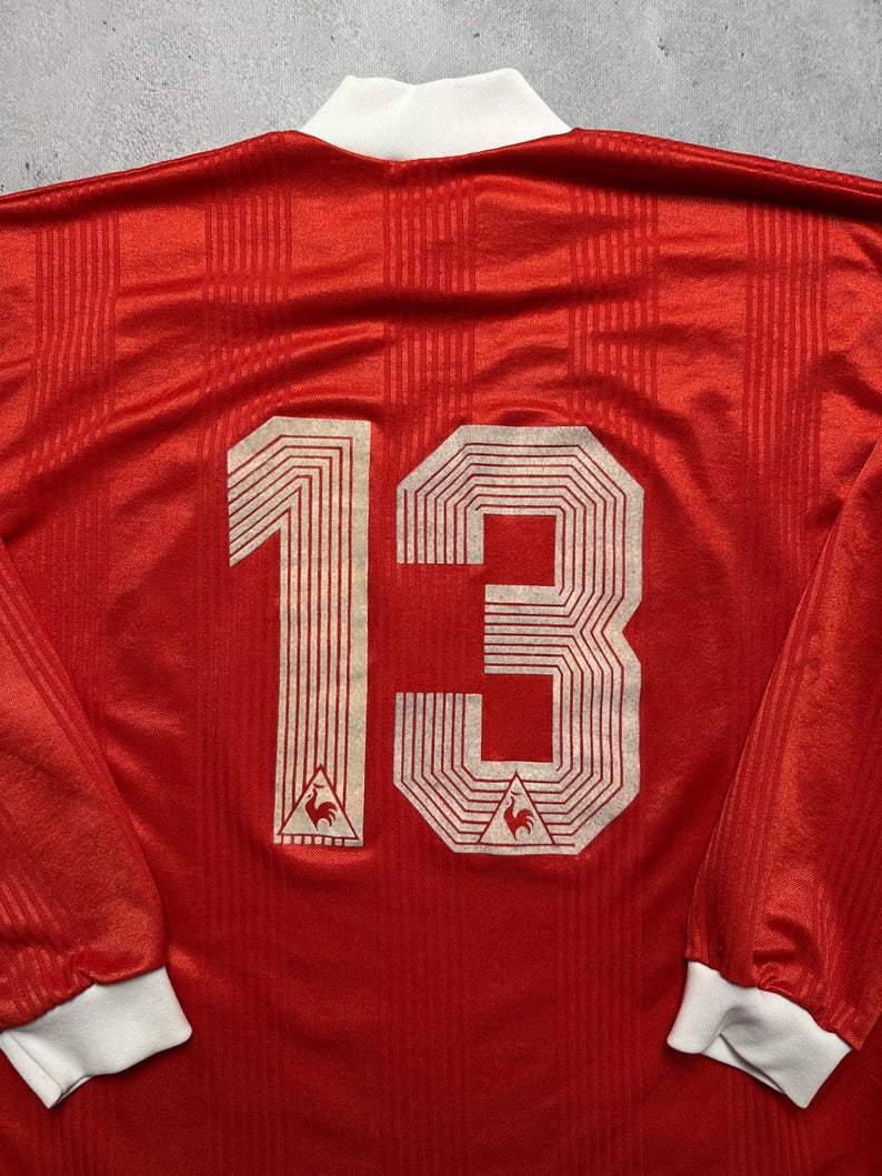 Puede incluir: Camiseta roja de manga larga con el n&uacute;mero 13 en blanco en la espalda. La camiseta tiene cuello y pu&ntilde;os blancos. Un peque&ntilde;o logotipo es visible debajo del n&uacute;mero. La tela tiene un sutil patr&oacute;n de rayas verticales.