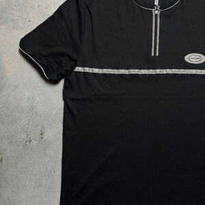 May include: Black short-sleeved t-shirt with a zipper at the neck and a silver stripe across the chest. The shirt has a small logo on the right side of the chest.