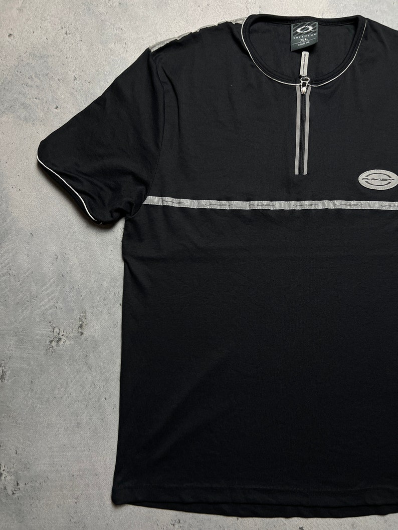 May include: Black short-sleeved t-shirt with a zipper at the neck and a silver stripe across the chest. The shirt has a small logo on the right side of the chest.