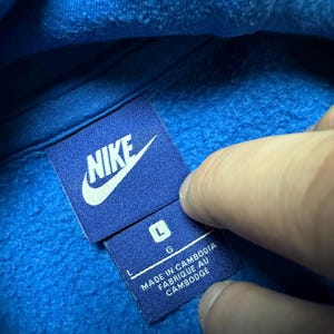 Peut inclure: Sweat &agrave; capuche bleu Nike avec un logo Nike blanc et une &eacute;tiquette indiquant "Fabriqu&eacute; au Cambodge" et "L G".