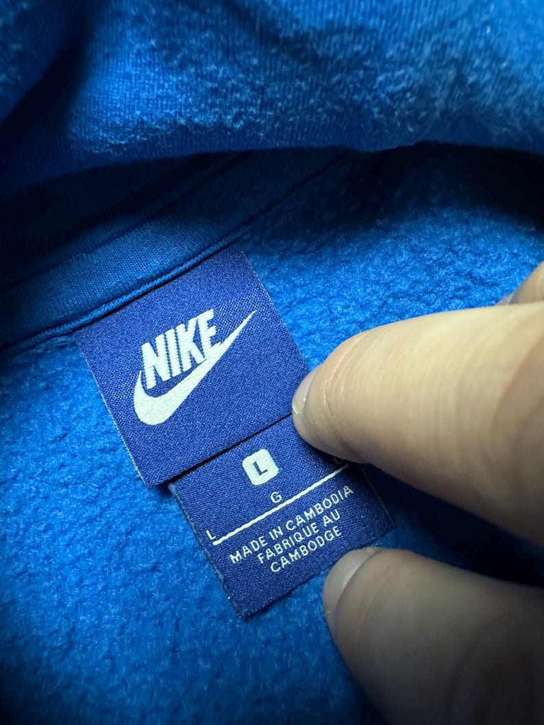Peut inclure: Sweat &agrave; capuche bleu Nike avec un logo Nike blanc et une &eacute;tiquette indiquant "Fabriqu&eacute; au Cambodge" et "L G".