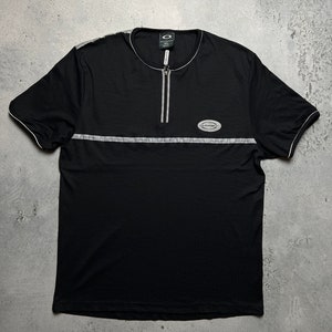May include: Black short-sleeved shirt with a zipper and a silver reflective stripe across the chest. The shirt has a small logo on the chest that says "Overkill" and a larger logo on the left sleeve that says "Overkill".