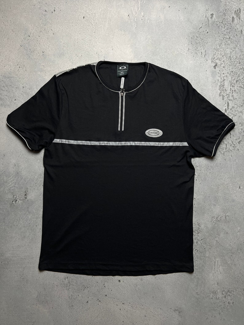 May include: Black short-sleeved shirt with a zipper and a silver reflective stripe across the chest. The shirt has a small logo on the chest that says "Overkill" and a larger logo on the left sleeve that says "Overkill".