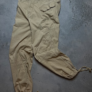 May include: Khaki-colored cargo pants with multiple pockets and drawstrings at the ankles. The pants feature the Nike logo and a button closure. The fabric appears to be a durable, woven material.