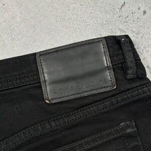 Pode incluir: Cal&ccedil;as de ganga pretas com um emblema retangular de couro preto no c&oacute;s. O emblema tem o texto Acne Studios gravado em relevo. As cal&ccedil;as incluem uma presilha para cinto e costuras vis&iacute;veis ao longo do tecido e do emblema.