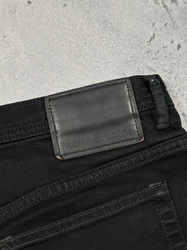 Pode incluir: Cal&ccedil;as de ganga pretas com um emblema retangular de couro preto no c&oacute;s. O emblema tem o texto Acne Studios gravado em relevo. As cal&ccedil;as incluem uma presilha para cinto e costuras vis&iacute;veis ao longo do tecido e do emblema.