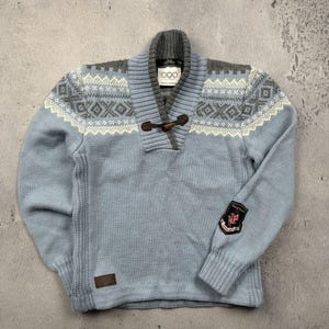 May include: A light blue sweater with a shawl collar and a toggle closure. The sweater features a gray and white geometric pattern across the chest and a patch on the left sleeve. The Olympic rings logo is visible on the collar.