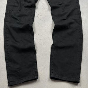Pode incluir: Um par de cal&ccedil;as de ganga pretas lisas com um design cl&aacute;ssico de cinco bolsos, fecho de bot&otilde;es e costuras vis&iacute;veis no mesmo tom ao longo das pernas. O tecido tem um acabamento mate com vincos subtis ao longo das pernas.