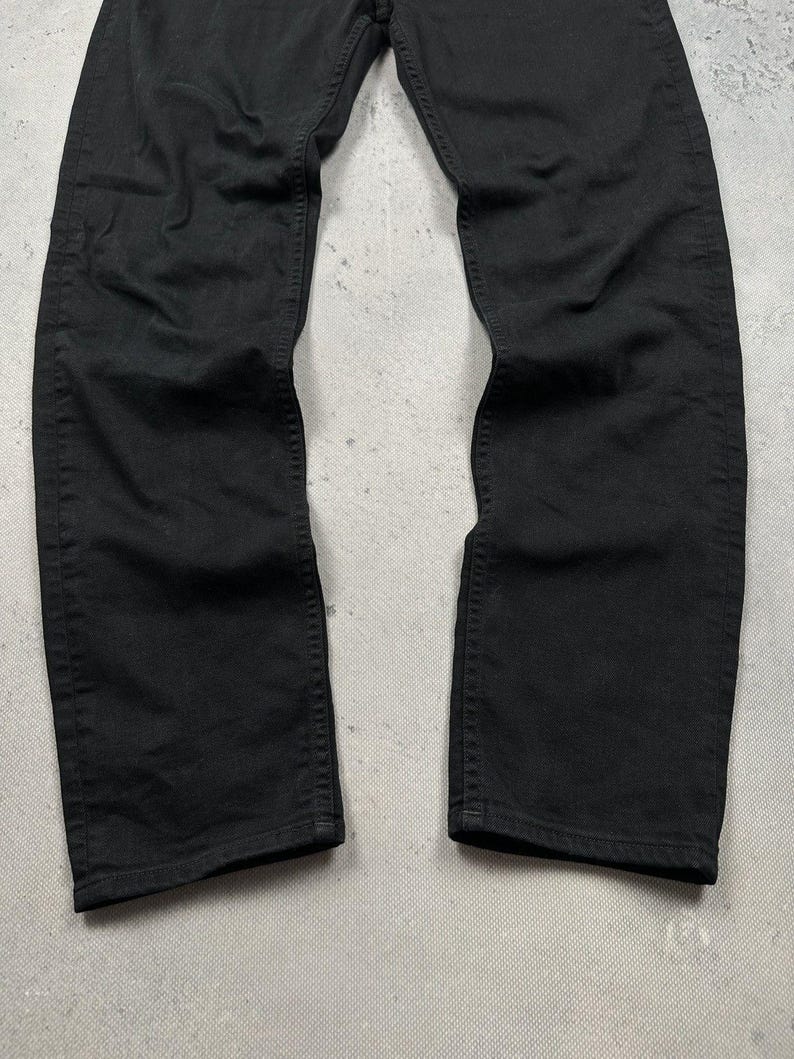 Pode incluir: Um par de cal&ccedil;as de ganga pretas lisas com um design cl&aacute;ssico de cinco bolsos, fecho de bot&otilde;es e costuras vis&iacute;veis no mesmo tom ao longo das pernas. O tecido tem um acabamento mate com vincos subtis ao longo das pernas.