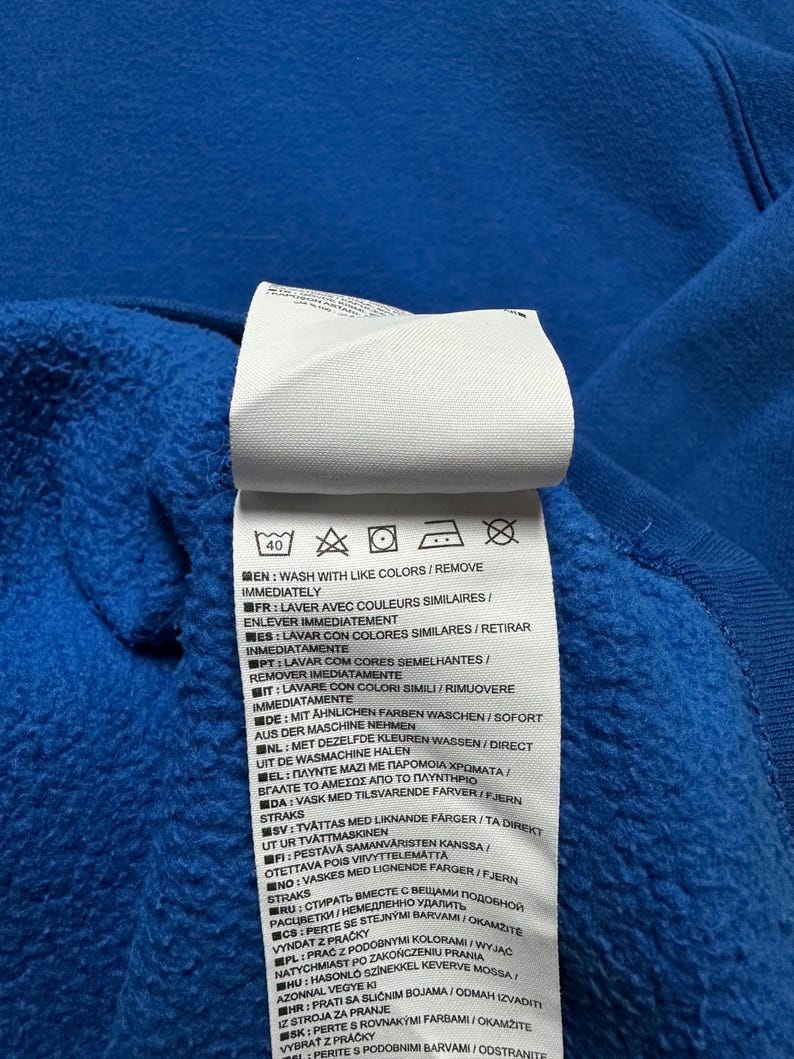 Peut inclure: Une &eacute;tiquette d'entretien blanche avec des instructions de lavage pour un v&ecirc;tement. L'&eacute;tiquette contient du texte dans plusieurs langues, notamment l'anglais, le fran&ccedil;ais, l'espagnol, l'italien, l'allemand, le n&eacute;erlandais, le portugais, le polonais et le su&eacute;dois. L'&eacute;tiquette indique de laver avec des couleurs similaires et de retirer imm&eacute;diatement.