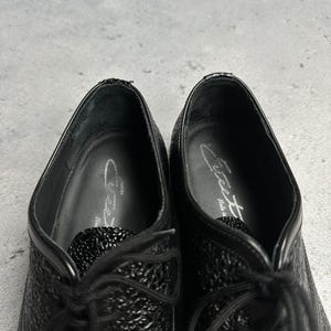 Może przedstawiać: Parafrazując, czarne sk&oacute;rzane buty wizytowe z teksturowanymi detalami. Buty mają sznurowany design i błyszczące wykończenie. Wnętrze but&oacute;w posiada nadrukowane logo. Buty są ustawione na szarym tle.