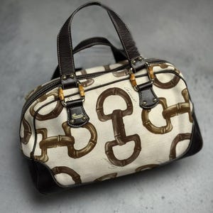 May include: A beige handbag with a brown equestrian-themed pattern. The bag features brown leather handles, trim, and accents, with bamboo-style hardware. The brand name "Gucci" is visible on the bag.