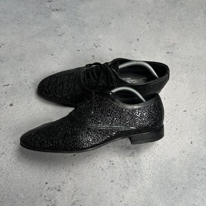 Może przedstawiać: Czarne sk&oacute;rzane buty wizytowe z teksturowanym wzorem. Buty mają klasyczny design sznurowany i niski obcas. Buty są prezentowane na szarej powierzchni.