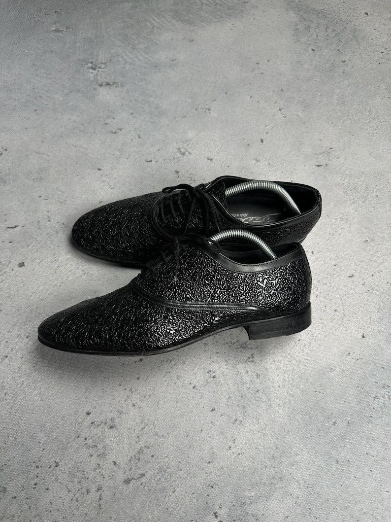 Może przedstawiać: Czarne sk&oacute;rzane buty wizytowe z teksturowanym wzorem. Buty mają klasyczny design sznurowany i niski obcas. Buty są prezentowane na szarej powierzchni.