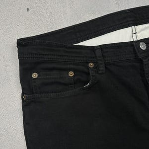 Pode incluir: Um par de cal&ccedil;as de ganga pretas com um design cl&aacute;ssico de cinco bolsos, fecho de bot&otilde;es, rebites met&aacute;licos em tom de cobre e um forro interior branco na cintura. O tecido tem uma textura de sarja vis&iacute;vel e costuras resistentes.
