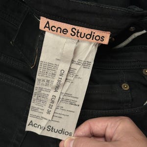 Pode incluir: Cal&ccedil;as de ganga pretas com uma etiqueta da marca Acne Studios em tom p&ecirc;ssego claro. Uma etiqueta de cuidados branca e comprida est&aacute; anexada, exibindo o texto Acne Studios, CN 195/90A e EUR 33 36, juntamente com detalhes da composi&ccedil;&atilde;o do tecido.