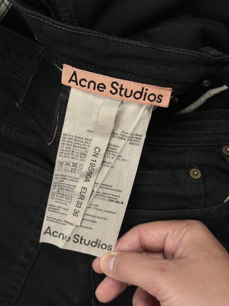 Pode incluir: Cal&ccedil;as de ganga pretas com uma etiqueta da marca Acne Studios em tom p&ecirc;ssego claro. Uma etiqueta de cuidados branca e comprida est&aacute; anexada, exibindo o texto Acne Studios, CN 195/90A e EUR 33 36, juntamente com detalhes da composi&ccedil;&atilde;o do tecido.
