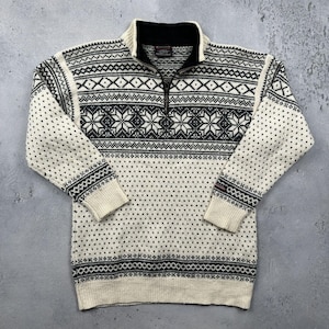 May include: Cream-colored sweater with a black and white geometric pattern. The sweater features a black collar, a quarter-zip closure, and ribbed cuffs and hem. The design includes diamond and snowflake motifs.