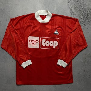 Puede incluir: Una camiseta de f&uacute;tbol roja de manga larga con cuello y pu&ntilde;os blancos. La camiseta presenta la palabra "Coop" en un rect&aacute;ngulo blanco y el logo "Le Coq Sportif". La camiseta tiene rayas verticales.