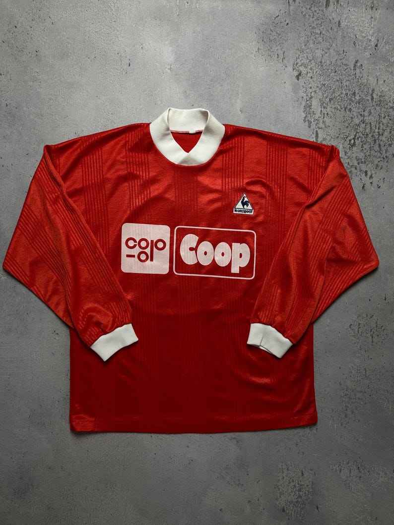 Puede incluir: Una camiseta de f&uacute;tbol roja de manga larga con cuello y pu&ntilde;os blancos. La camiseta presenta la palabra "Coop" en un rect&aacute;ngulo blanco y el logo "Le Coq Sportif". La camiseta tiene rayas verticales.