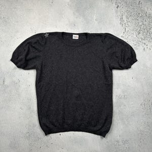 Puede incluir: Un suéter gris oscuro de manga corta con cuello redondo y mangas abullonadas. El suéter tiene una pequeña etiqueta en el cuello y un detalle decorativo en el hombro. La prenda está hecha de un material suave y texturizado.