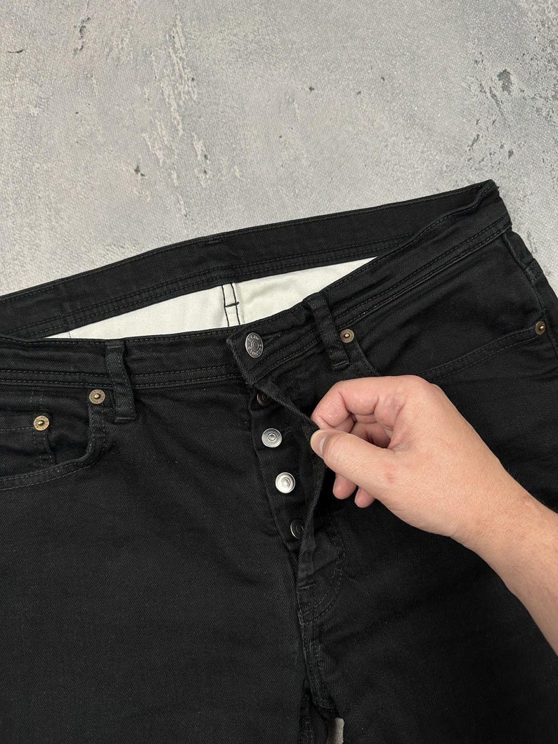Pode incluir: Cal&ccedil;as de ganga pretas com fecho de bot&otilde;es em metal prateado, c&oacute;s com presilhas para cinto e um pequeno bolso para moedas no lado frontal direito. O forro interior do c&oacute;s &eacute; de tecido branco com costuras vis&iacute;veis.