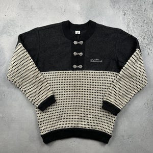 May include: A vintage black and cream-colored sweater with a unique design. The upper portion is solid black, while the lower section and sleeves feature a cream-colored pattern with black dots. The sweater has decorative clasps and the word "Telemark" embroidered on the front.