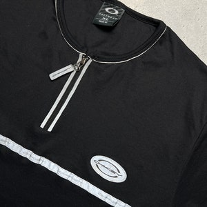 May include: Black long-sleeve shirt with a white zipper and white trim. The shirt has a white oval logo with the word "Oakley" in black text.