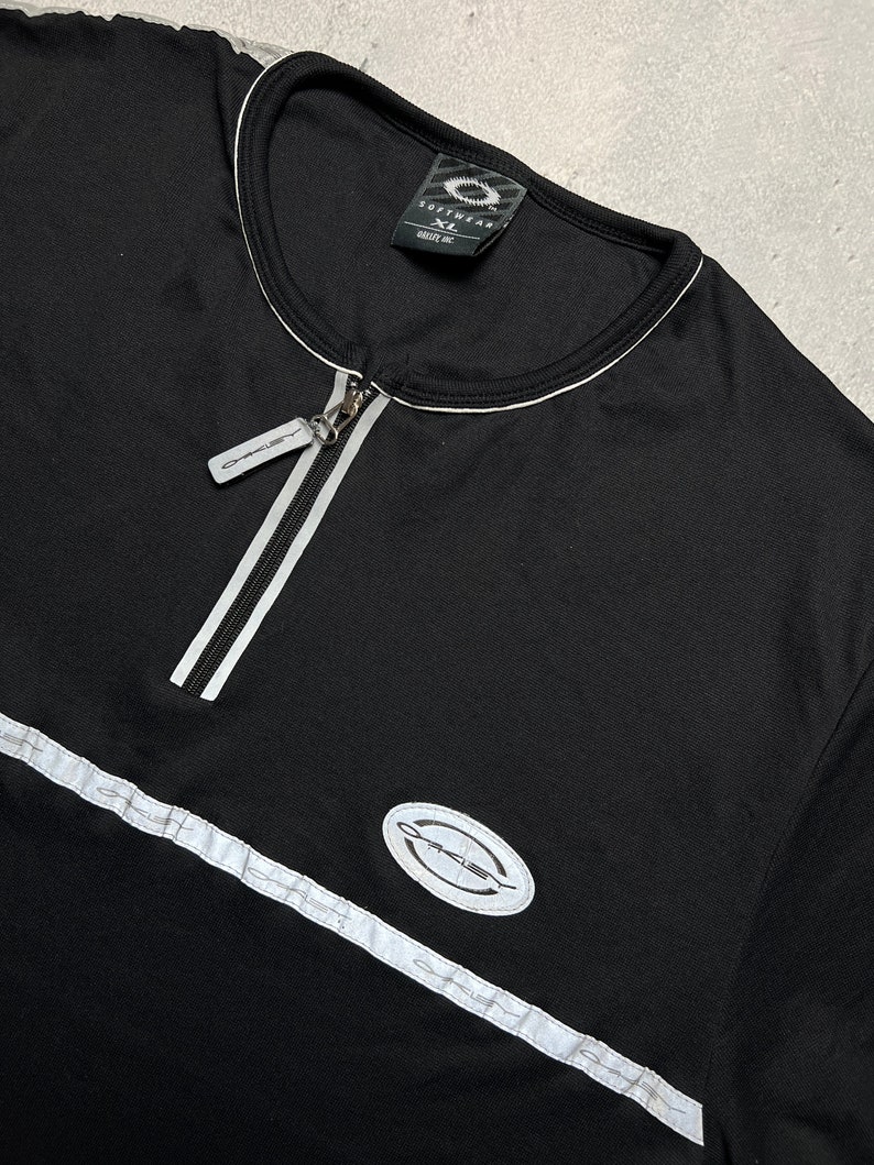 May include: Black long-sleeve shirt with a white zipper and white trim. The shirt has a white oval logo with the word "Oakley" in black text.