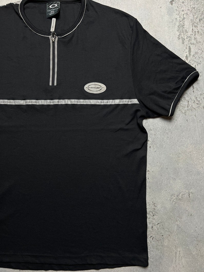 May include: Black short-sleeved t-shirt with a zipper and a silver reflective stripe across the chest. The shirt has a small oval logo with the text "Essential" on it.