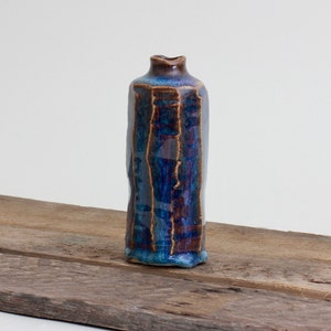 May include: A handmade ceramic vase with a rectangular shape and a narrow opening. The vase features a glossy glaze in shades of blue, brown, and teal. The vase is approximately 8 inches tall and sits on a wooden surface.