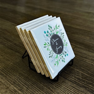 May include: Set of six white ceramic coasters with a floral design and the text "Shawna & Jeremy" in a black circle. The coasters are displayed on a black metal stand.