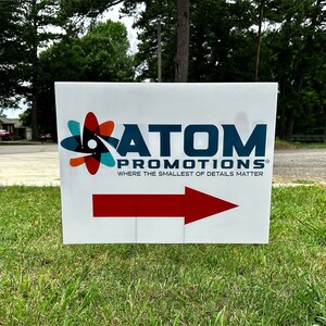 May include: White sign with a red arrow pointing to the right. The sign has a blue and white logo with the text "ATOM PROMOTIONS®" and the tagline "WHERE THE SMALLEST OF DETAILS MATTER".