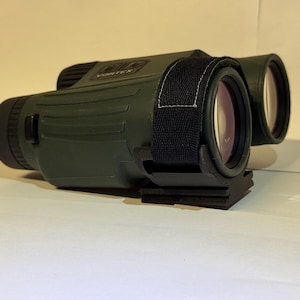 May include: Dark green binoculars with black accents and a black strap. The binoculars have a textured surface and a black base. The lenses are visible and have a reddish tint. The word "VORTEX" is visible on the top.