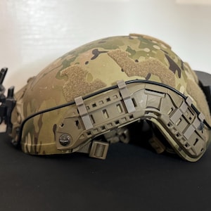 Puede incluir: Un casco con patrón de camuflaje con varios accesorios, incluyendo un sistema de rieles y un dispositivo negro en el lateral. El casco presenta tonos beige y verdes, con una visera oscura y herrajes de montaje.
