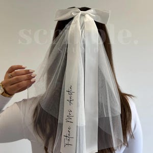 Pode incluir: Um véu de noiva branco com um laço grande no topo. O véu tem um design transparente e em camadas e apresenta o texto "Future Mrs Austin" impresso em escrita preta em uma fita branca.