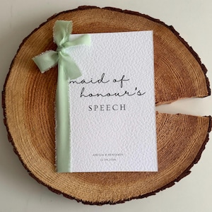 May include: A white textured card with the words "maid of honour's SPEECH" in black script, tied with a light green ribbon. The card rests on a wooden slice, with the names Amelia & Benjamin and the date 22.08.2026 printed at the bottom.