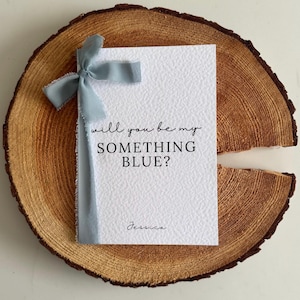 Könnte beinhalten: Eine weiße Karte mit dem Text "will you be my SOMETHING BLUE?" und dem Namen "Jessica" in Schreibschrift. Ein hellblaues Band ist um die Karte gebunden. Die Karte liegt auf einer Holzscheibe.