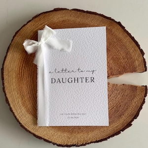 May include: A white card with the text "a letter to my DAUGHTER" and "ON YOUR WEDDING DAY 22.08.2026" printed in black. The card is tied with a white ribbon and rests on a wooden slice.
