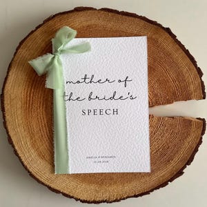 Könnte beinhalten: Eine weiße, strukturierte Karte mit dem Text "mother of the bride's SPEECH" in schwarzer Schrift. Ein hellgrünes Band ist um die Karte gebunden. Die Karte liegt auf einer runden Holzscheibe.