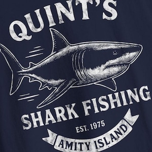 Op de afbeelding: Marineblauwe graphic tee met een witte haai-illustratie. De tekst luidt "QUINT'S SHARK FISHING" erboven en "AMITY ISLAND" eronder, met "EST. 1975" in het midden. Het ontwerp heeft een verweerde, vintage uitstraling.