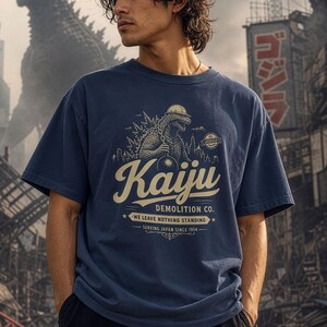 May include: A navy blue t-shirt featuring a graphic of a monster wearing a hard hat and holding a wrecking ball. The text reads Kaiju Demolition Co. We Leave Nothing Standing. Serving Japan Since 1954. The design is printed in a cream color.